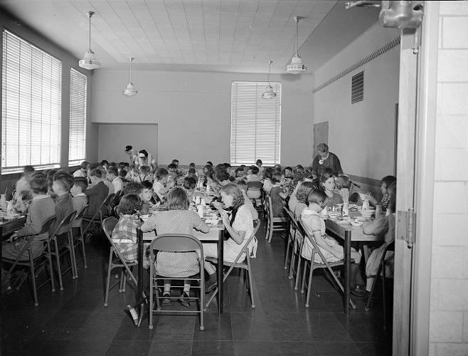 Children sit at rows of tables in a small room eating lunch. 