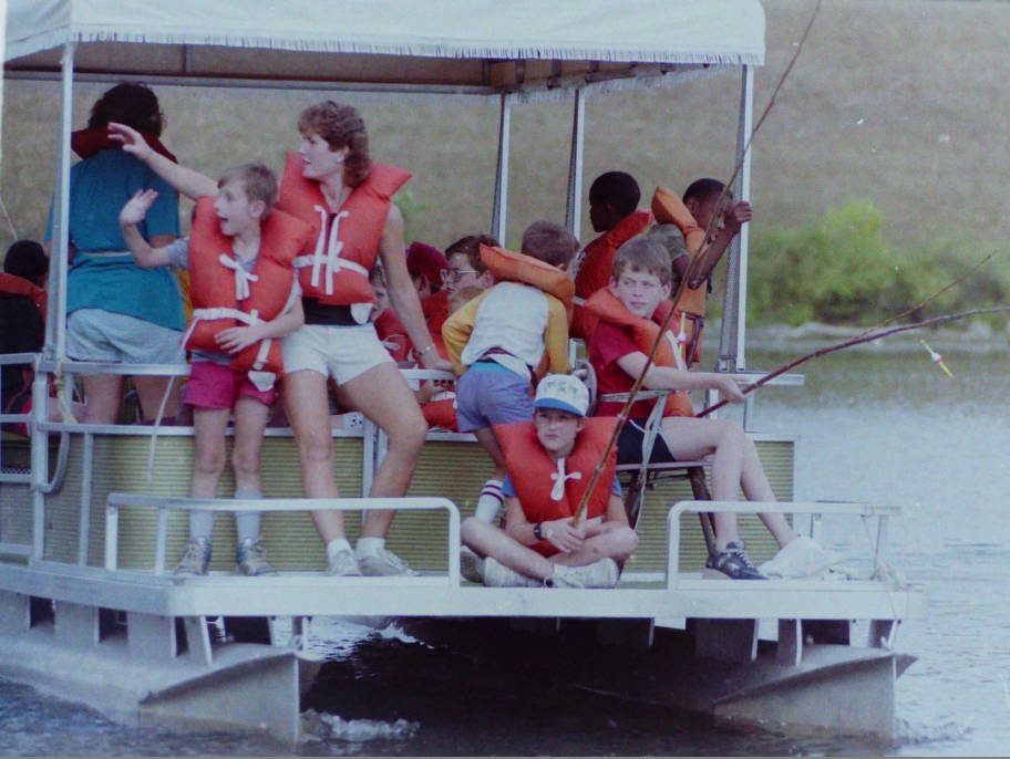 A group of people, mainly children, are on a pontoon boat. They are wearing life vests and some are holding fishing rods.
