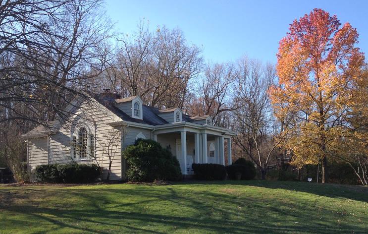 A one-story gray house with a gabled roof and a portico sits on a sloping lawn surrounded by trees.
