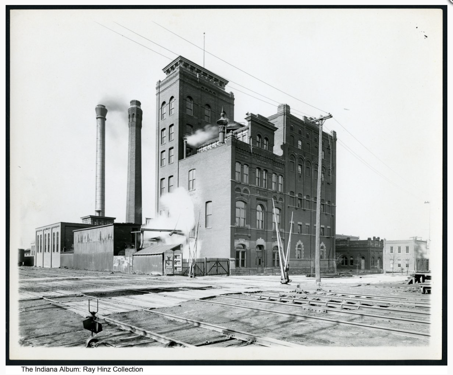 A large, multi-level, brick, industrial collection of attached building with two tall smokestacks is shown. There are railroad tracks in front.