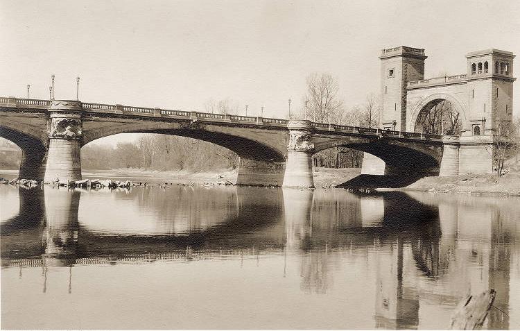 A low arch bridge with decorative piers spans the river and ends in a stone arch flanked by square turrets.