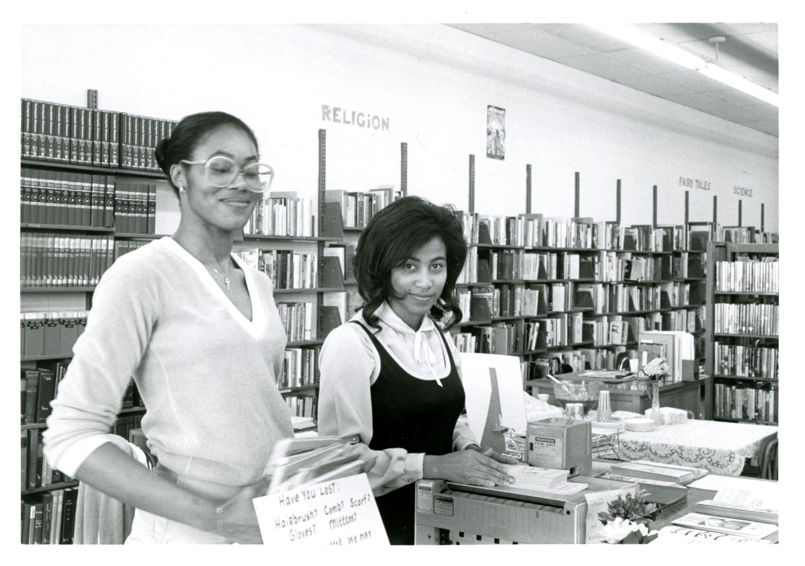 Two women stand in a library.