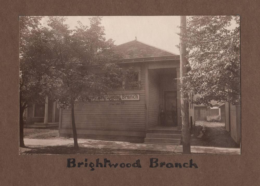 Front of a small building. A sign affixed to the front reads "Brightwood Branch." Children stand at the door.