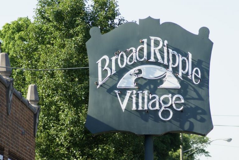 A sign mounted on a post says "Broad Ripple Village". There is an image of a duck on it.
