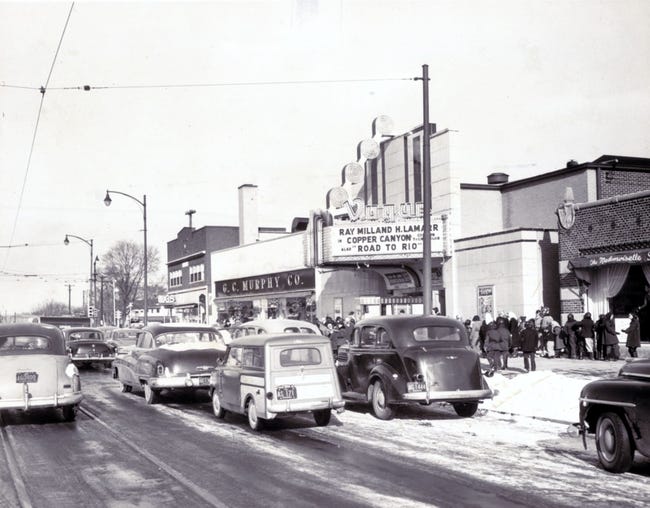Several cars are on a three-lane road that runs in front of a length of storefronts. Near the middle of the row is a theatre with a marquee that reads "Ray Milland and H. Lamar in Copper Canyon and Road to Rio"