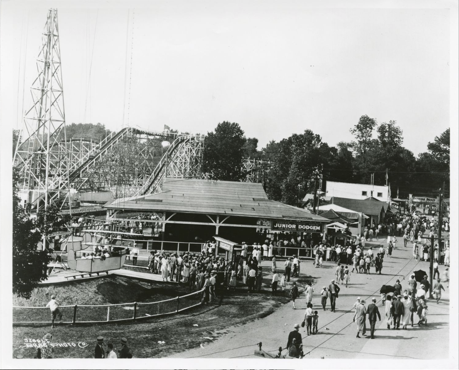 A view of many people on the grounds of an amusement park. A low covered building in the center has a sign for "Junior Dodgem". There is a large roller coaster in the background.