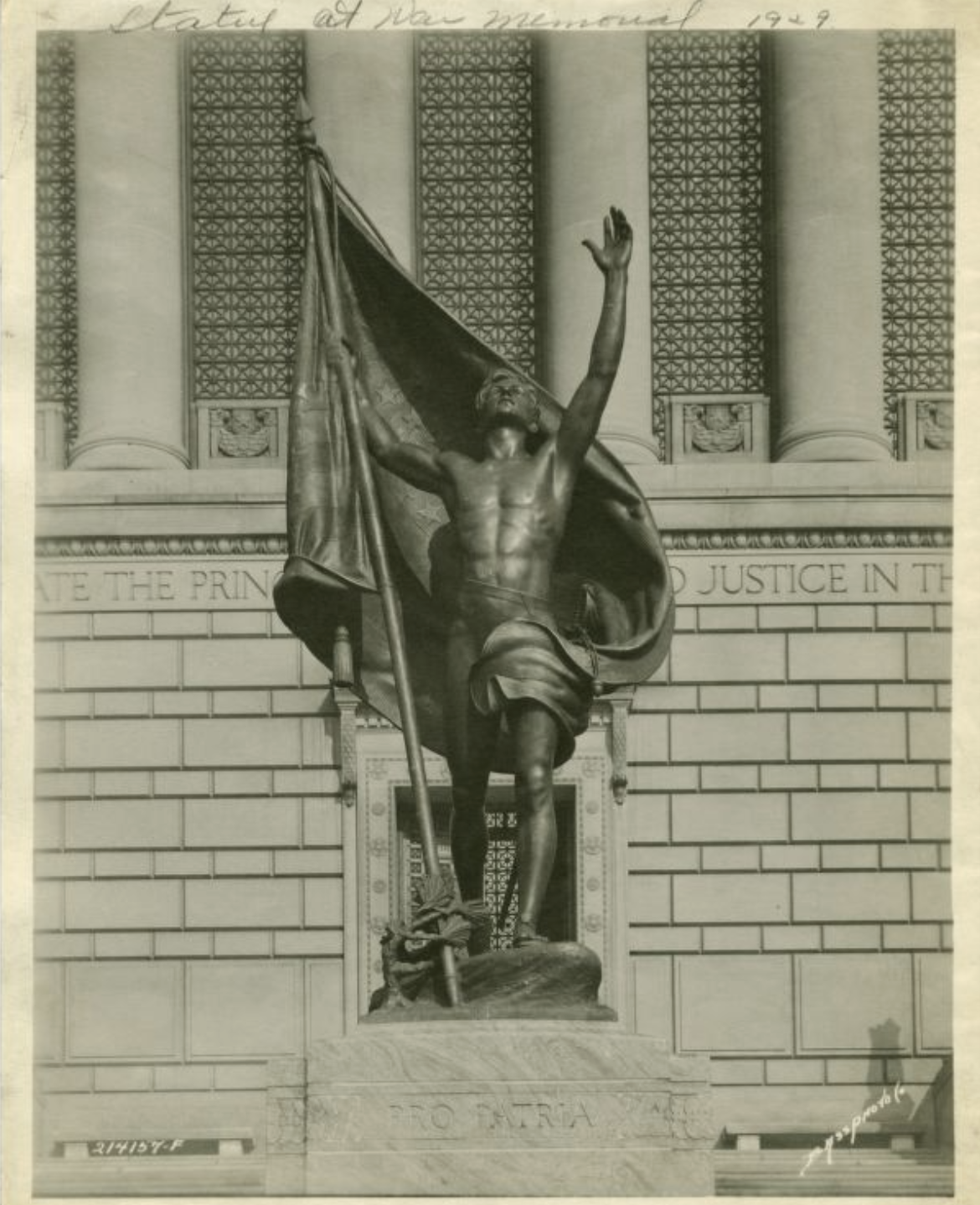 A bronze statue in front of a building. The statue features a shirtless man with his arms raised up. The is holding a flag in one hand.