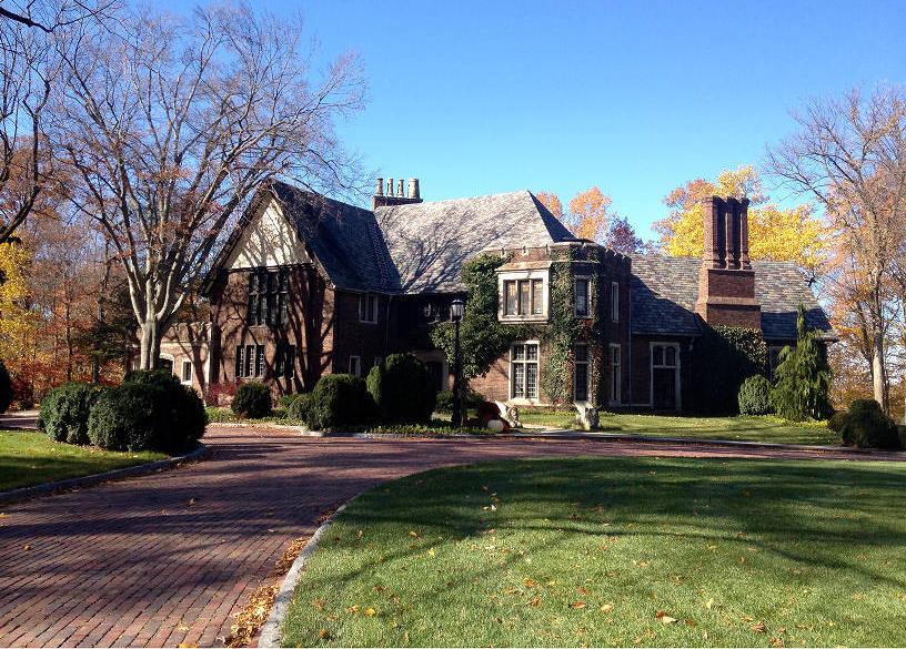 A wide brick circle drive way leads up to a large, two-story brick home. The English country manor-style house has a gable roof and a crenellated tower structure in the front corner.
