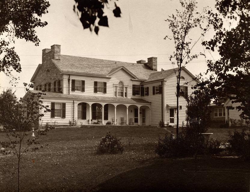 A large, white clapboard house sits on a large lawn. It has a gable roof with twin stone chimneys at each end. There is a long covered front porch, and the windows have dark shutters.