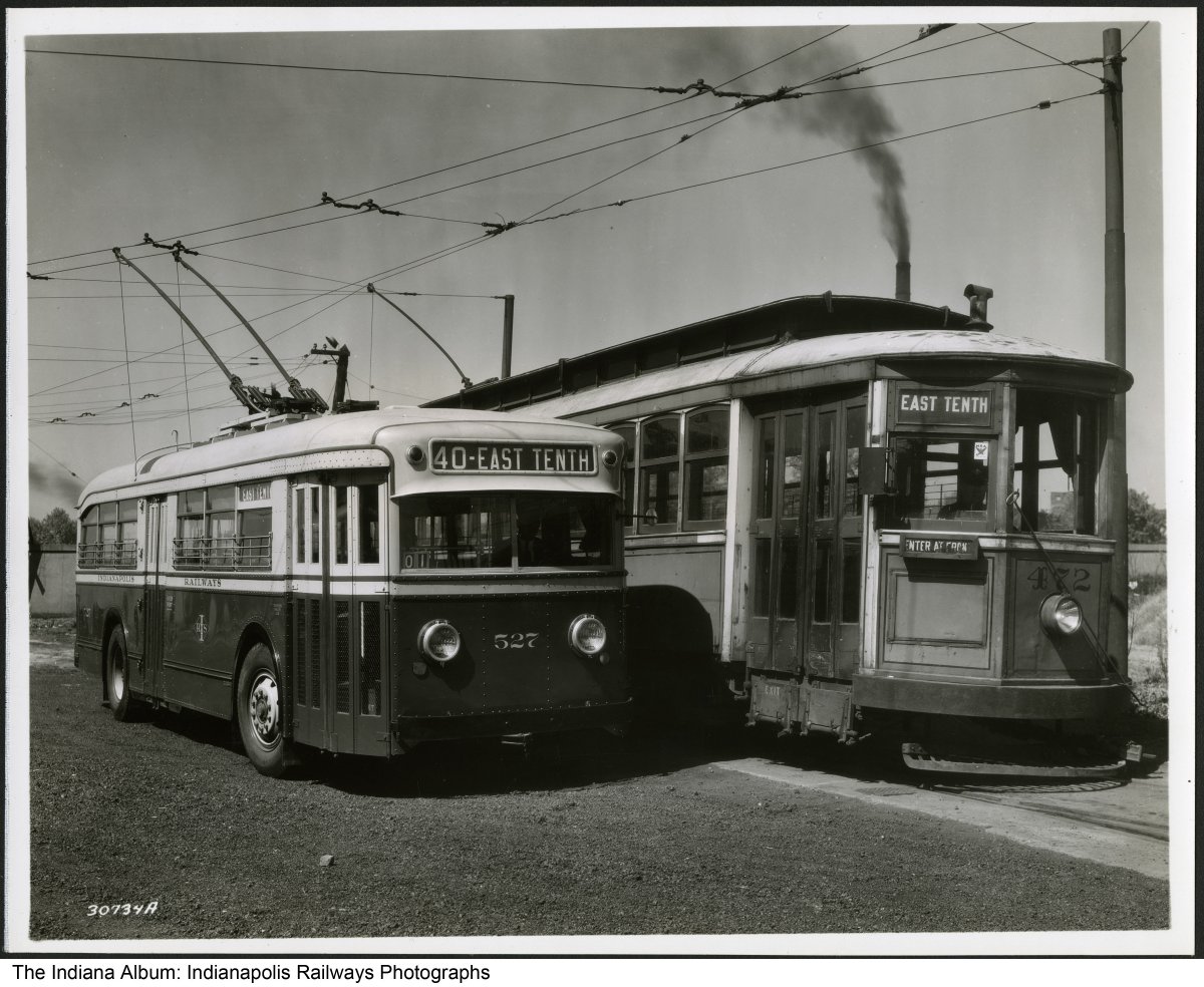 A larger streetcar is shown next to the small bus. Both have electrical conductors on the roof which reach up to connect with the overhead powerlines.