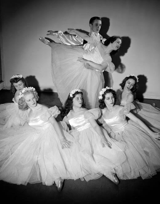 A male dancer supports a woman in an arabesque pose while five other dancers are seated in a semi-circle in front of them. The women all wear tulle tutus with satin bodices.