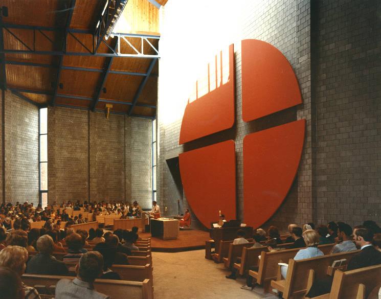 Interior view of a high-ceilinged room shows people sitting on wooden pews around an alter.