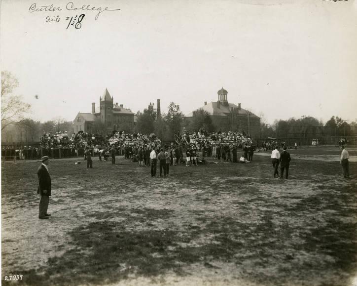 Many people are in the bleachers on the side of an open baseball field. Several other people are standing around in the outfield.