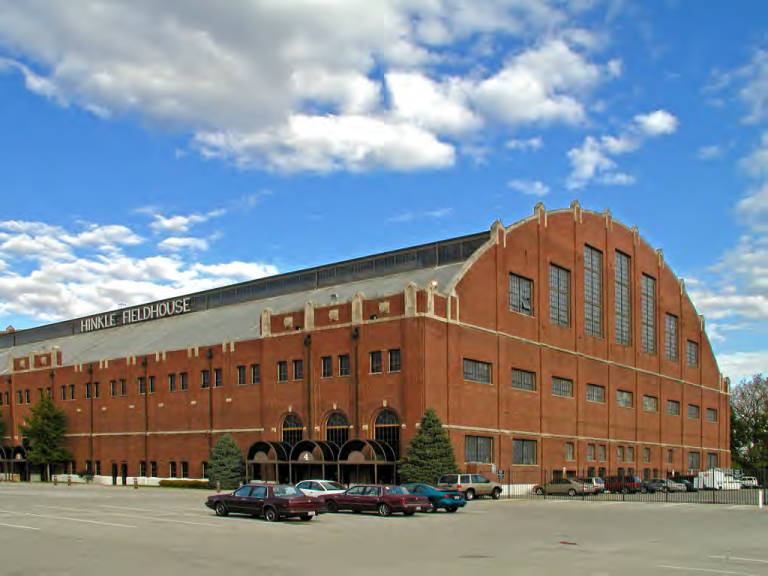 The large fieldhouse is shaped like an airplane hangar. The walls are all brick with mullioned windows of various sizes. The double-pitched roof has a monitor, which is a low, raised structure that runs down the spine of the roof and is lined on each side with windows.