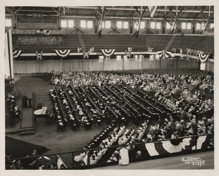 On the floor of the fieldhouse, rows of people are seated in front of a dais with a man standing at a podium.