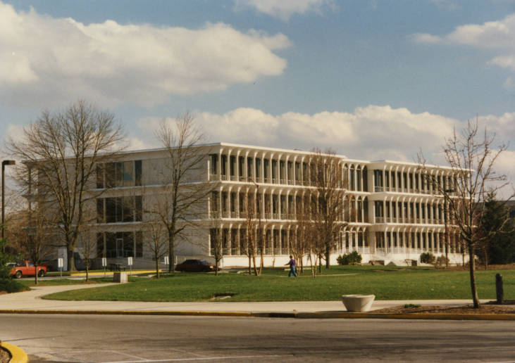 The building is composed of three, long rectangular tiers. The front of each tier is a long row of white columns, set close together and framing arched windows.
