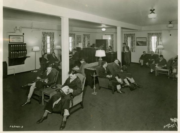 A group of men in pajamas and robes lounge in chairs in a large open room. 