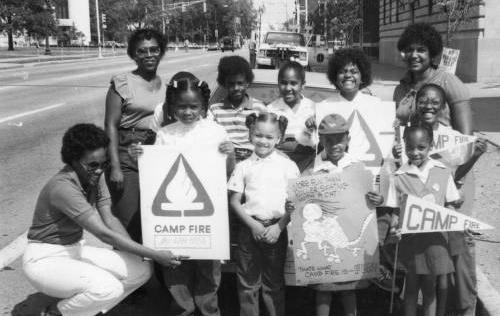 A group of children with three adults stand holding various Camp Fire signs.