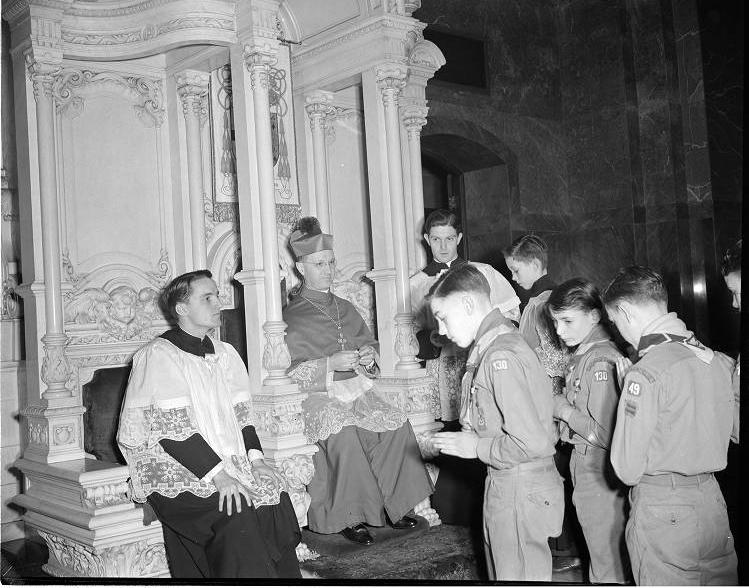 The bishop is seated in an ornate, Grecian revival structure of stone with columns. There are acolytes seated at his sides, and Boy Scouts stand in front of him.