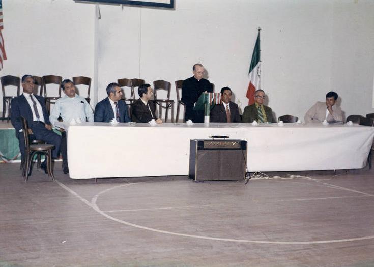 A line of men are seated behind a long table. A priest stands in the middle at a lectern.