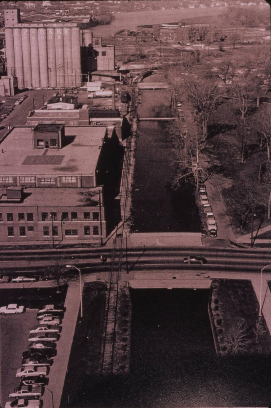 Aerial view showing the central canal with a road crossing the canal. Buildings are on one side of the canal and a grassy space is on the other. 