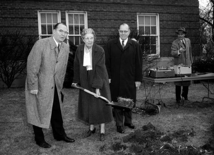 Three people stand outside a building. The woman in the center is holding a shovel with a piece of sod on it. There is a man standing at a table behind the group.