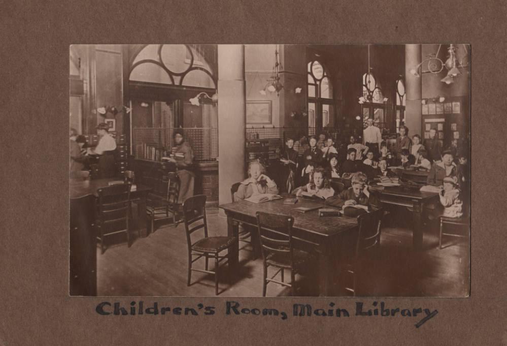 A large room is filled with tables. Children stand around the room and sit at the tables with open books.