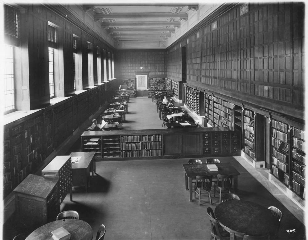 A view down a long hall way. Bookshelves line the walls. The hall is filled with more bookshelves and tables. Several people sit at the tables.