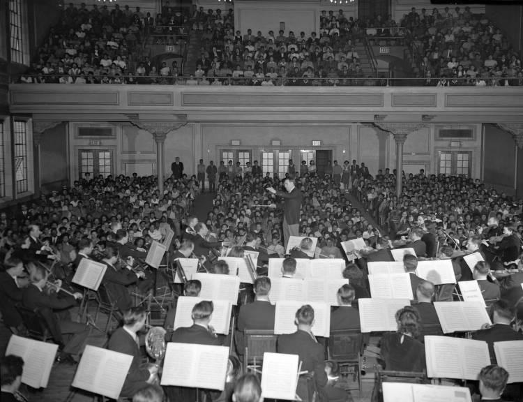 A large audience are in seats on the floor and balcony of an auditorium watching the symphony.