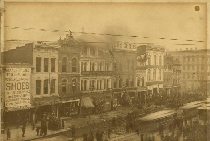 Smoke rises from the middle building in a row of false-fronted, old-fashioned storefronts. Several people are in the streets and firefighters climb ladders leaning against the front of the building.