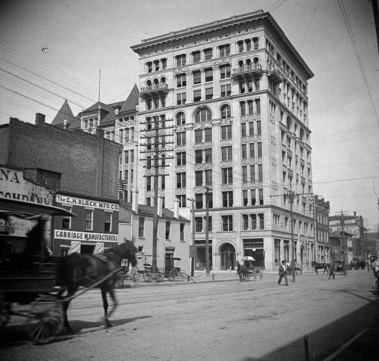 A three story brick building stands amid a long row of buildings facing a street with horse-drawn carriages on it.