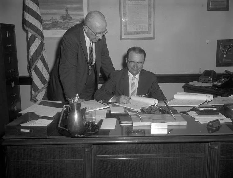 Two men are behind a desk. One is standing and leaning over the seated man as he signs a document. There is an American flag furled in the corner.