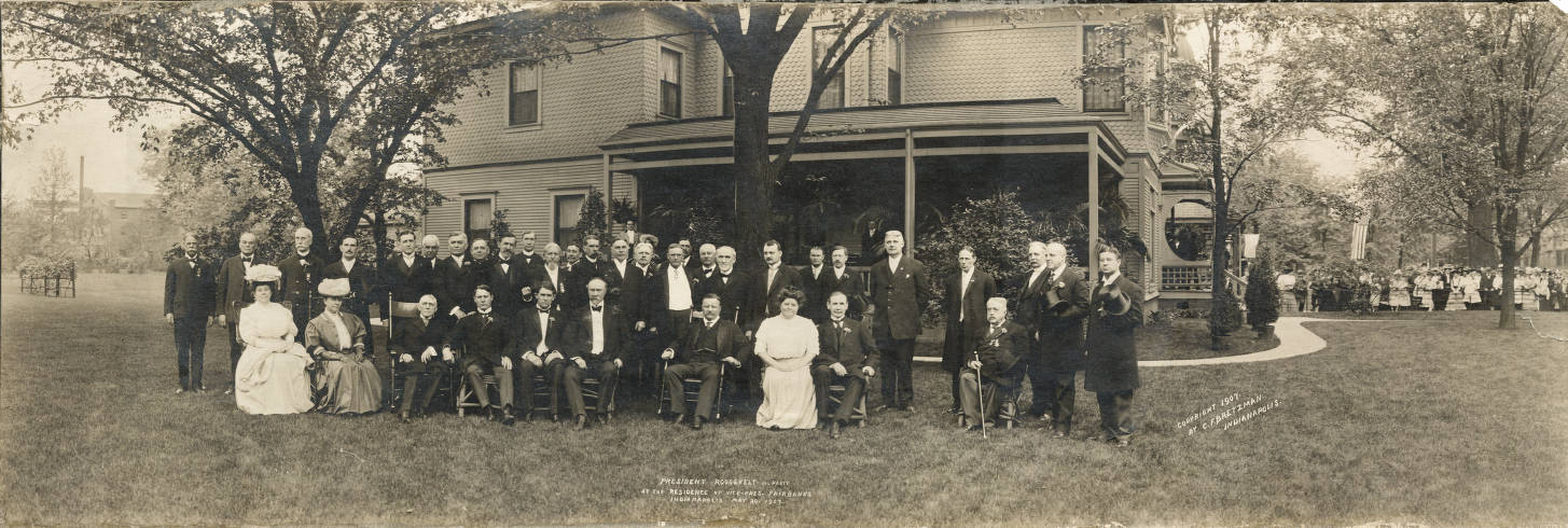 Two rows of people are in the side yard of a Victorian house. Beyond the house, a large crowd is gathered. The president is seated in the center of the front row.