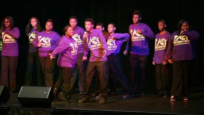 A group of people in purple shirts ( with "ACT" on the front) are on a stage. A young man in the middle is holding a microphone.