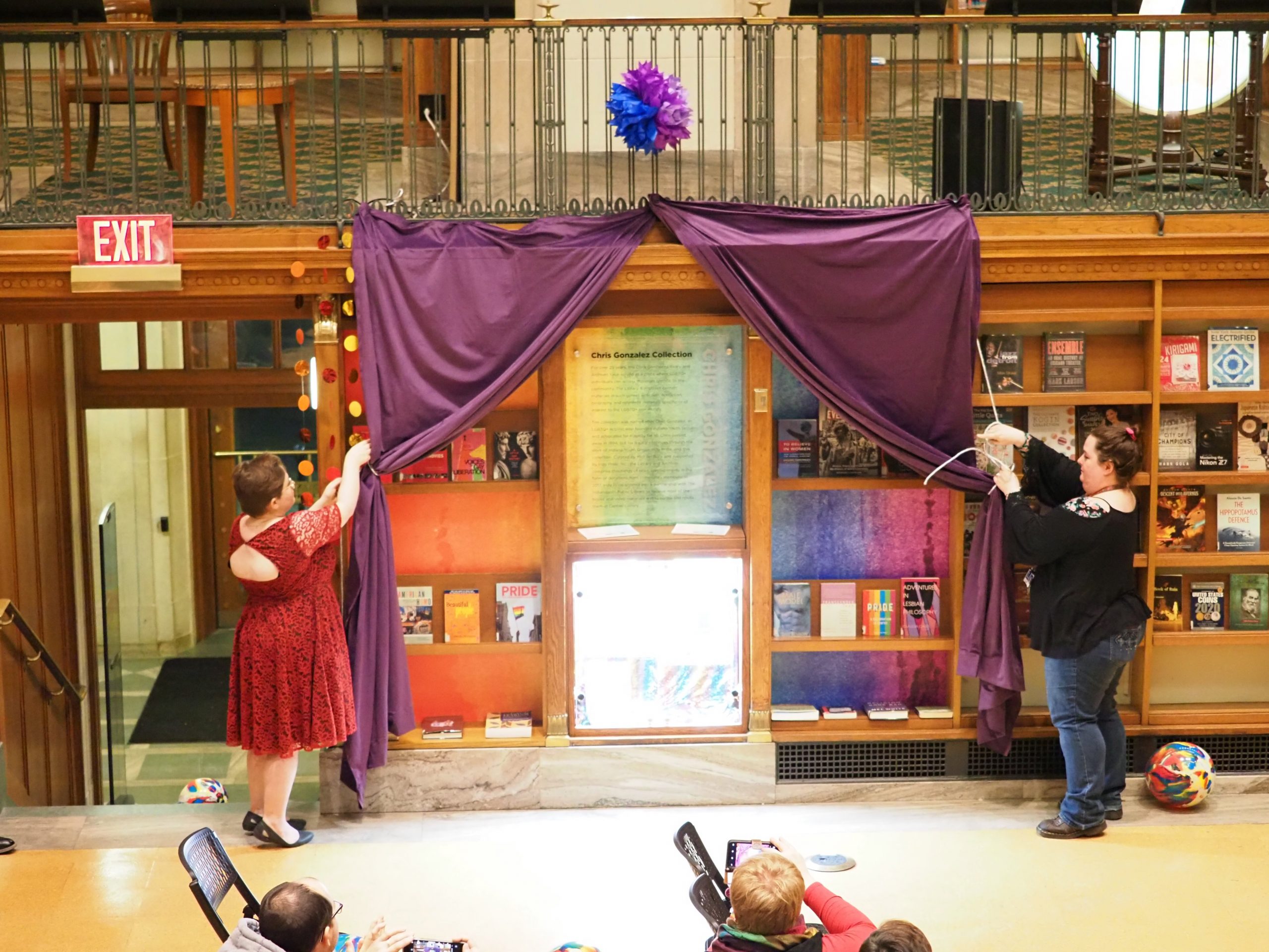Two women draw a purple curtain back to reveal a bookshelf display.
