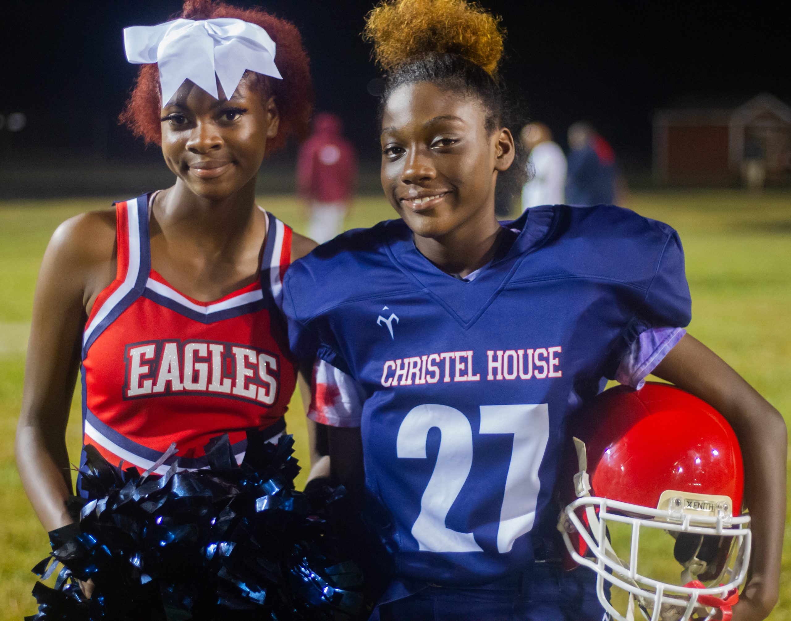 Two girls pose on a football field. One girl is dressed as a cheerleader and the other girl is in a football uniform.