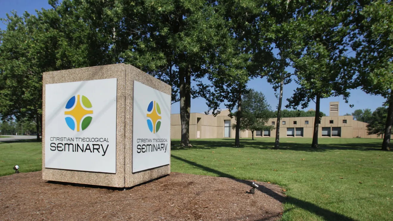 A stone cube-shaped sign stands in front of a long, low, modern sandstone building. Two sides of the sign are visible and each has "Christian Theological Seminary" on a white plaque attached to the face of the stone. On each is a circle around a stylized cross.