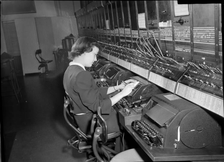 A woman sits in front of a switchboard. She is taking down a note. 