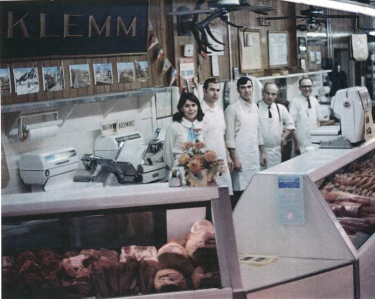 Five people wearing white aprons stand in a line behind a meat counter.