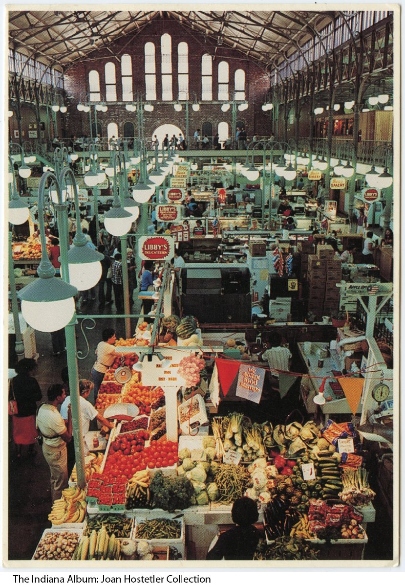 A produce stall is in the foreground. Rows of vendor stalls stretch beyond. Each stall has a street light with double, hanging globe lights suspended from it.