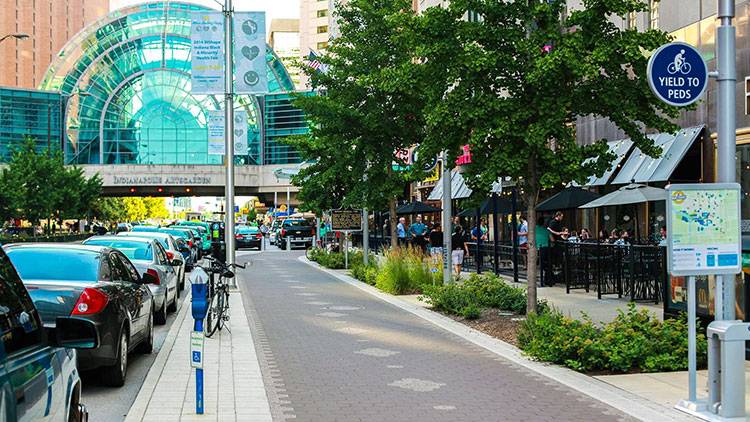 Street-level view of the Indianapolis Cultural Trail. The Circle Center Mall is on the far left. Shops and restaurants line the right side, and the glass dome of the Arts Garden spans the street in the background.
