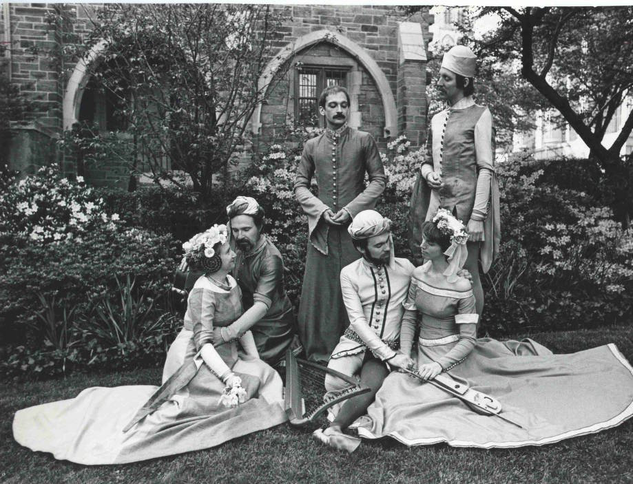 Six costumed people strike various poses on the lawn in front of a stone building.