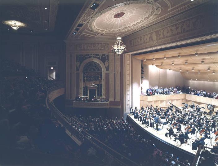 A view from the balconies shows the orchestra on the stage playing to a full audience.