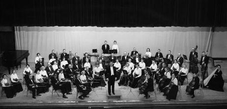 The conductor stands at center front of the stage, while the musicians are in semi-circulars rows behind.