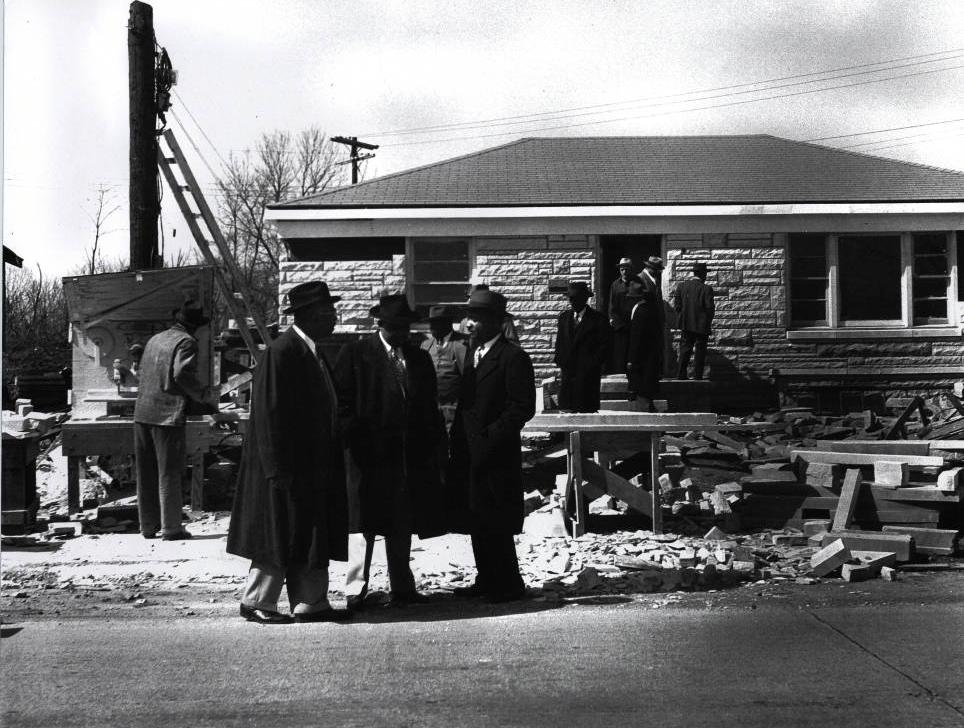 Several men stand amid construction materials in front of a stone-clad ranch-style house.