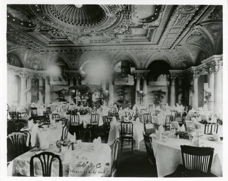 An elegant dining room with several tables covered in white tablecloths has an elaborately decorated coffer ceiling. The ceiling contains intricately carved panels. The walls of the room are composed of gothic arches supported by columns topped with decorative pediments.