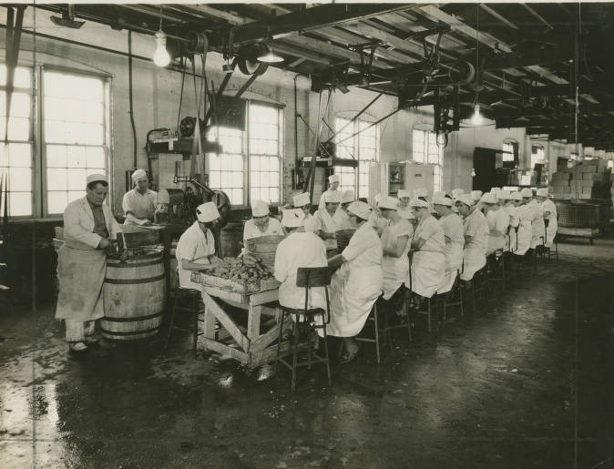 Women in white uniforms sit on each side of a long table in an industrial-looking room.