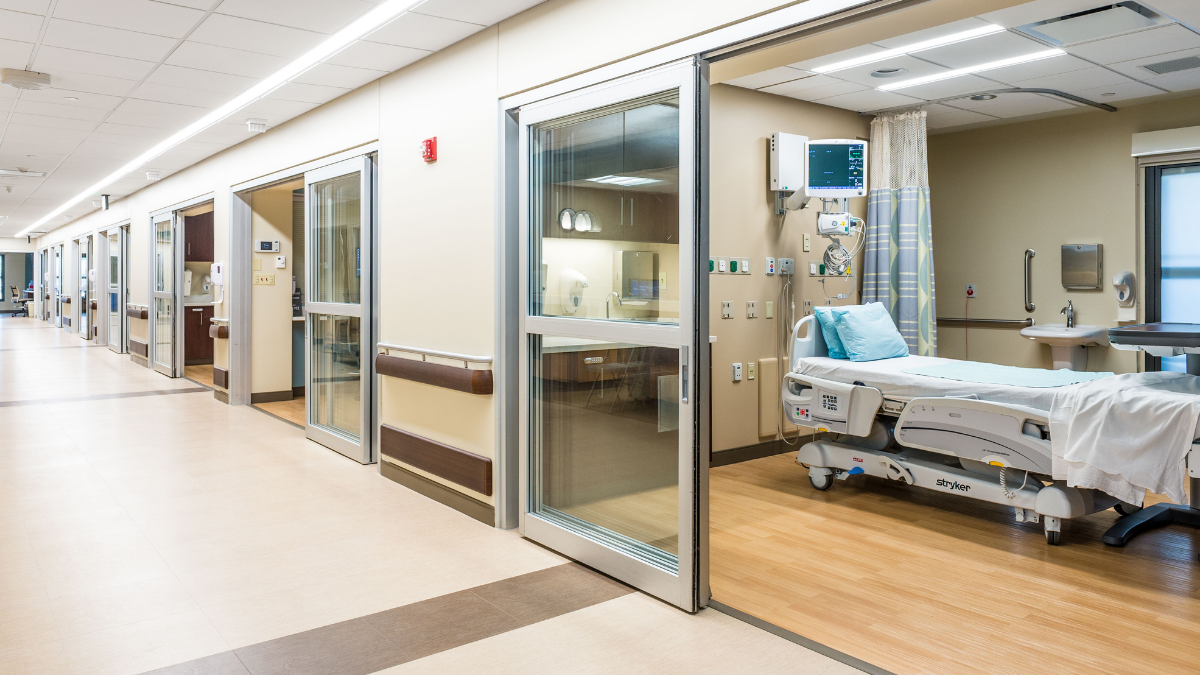 A hospital corridor is lined with glassed-front examination rooms.