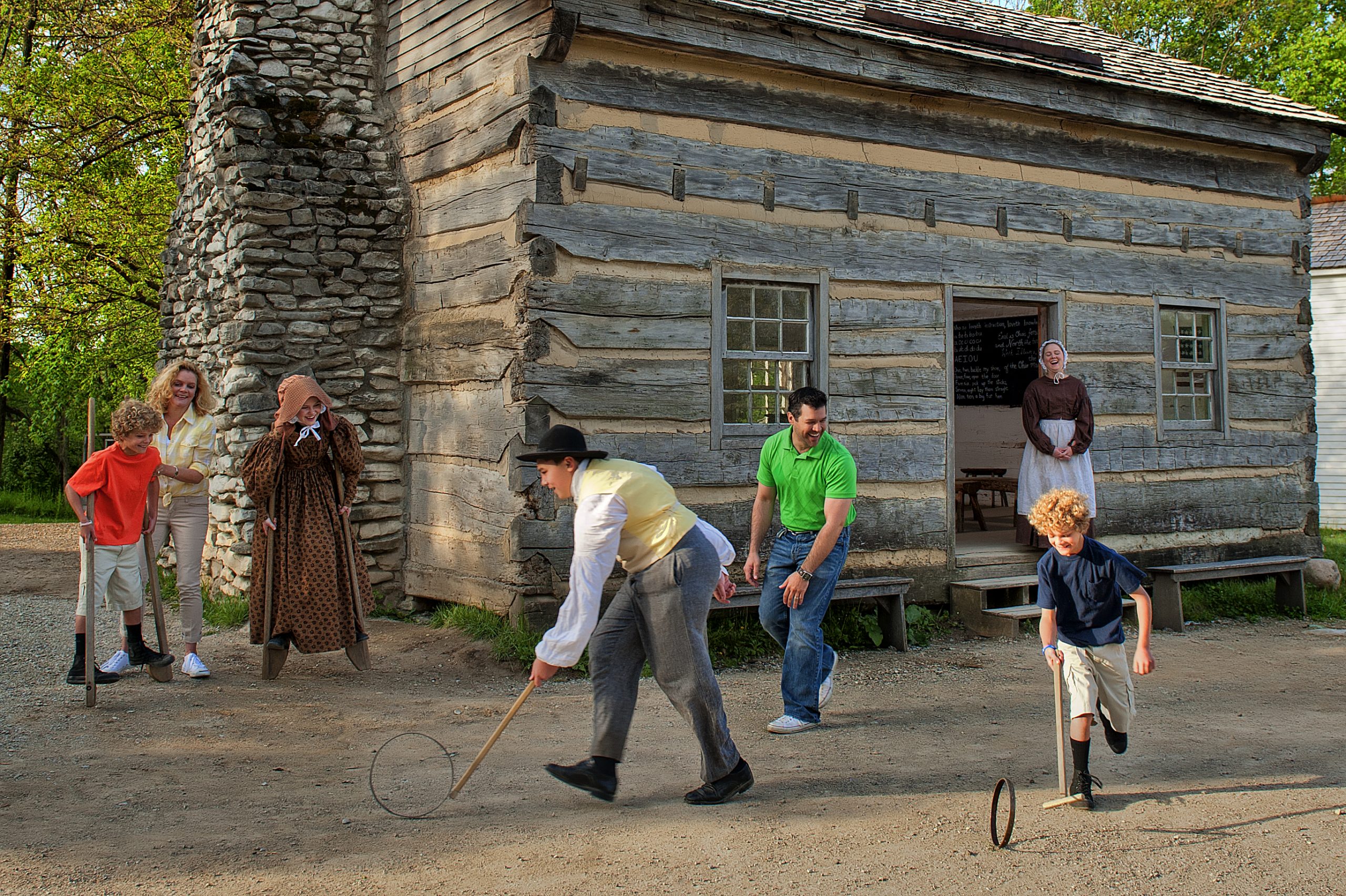 Three interpreters interact with a family. One interpreter shows a man and boy how to play a game with a hoop and stick. Another interpreter shows a child and woman how to use stilts. The other interpreter stands on the steps of the entrance to a log cabin.