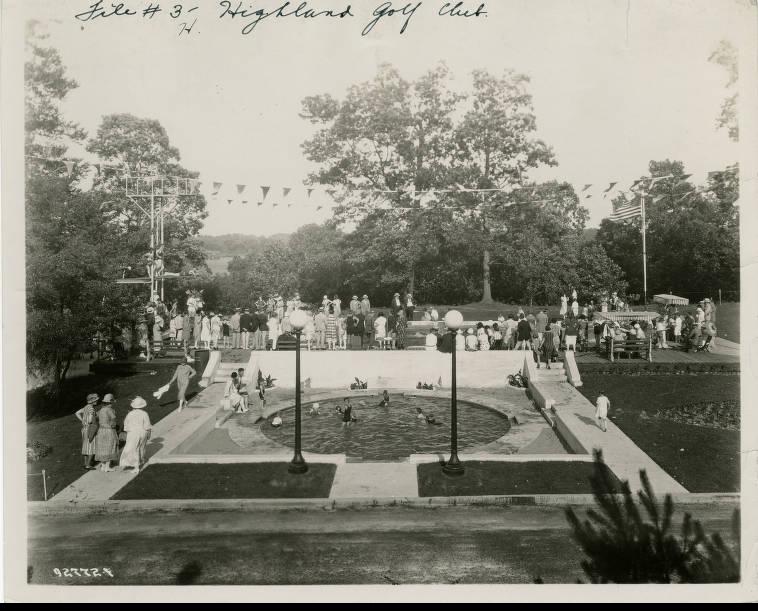 A shallow, circular swimming pool is in the foreground of this photo of the Highland Country Club, 1925. Several people stand around the patio behind the pool and many bathers are in it.